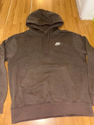 Brun hoodie från Nike - Säljer en brun hoodie från Nike med klassisk vit logga på bröstet. Hoodien har huva med dragsko och en stor magficka framtill. Tillverkad i mjukt bomullsmaterial och har ribbade muddar vid ärmslut och nederkant. Perfekt för en avslappnad och sportig stil.