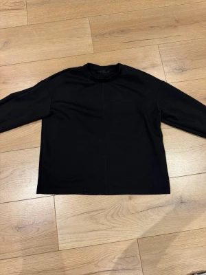 Svart långärmad tröja - En enkel svart långärmad tröja med rak passform och rund hals. Tröjan har en diskret söm mitt fram och är tillverkad i ett mjukt material som känns skönt mot huden. Perfekt att matcha med jeans eller kjol för en clean look.