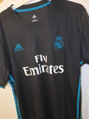 Real Madrid bortatröja Adidas svart - Snygg svart Real Madrid bortatröja från Adidas med blå detaljer och Fly Emirates-tryck på bröstet. Tröjan har klubbmärket och Adidas-loggan framtill samt rund hals och korta ärmar. Perfekt för fotbollsträning eller att bära som supporter.