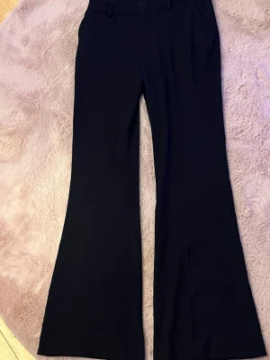 Svarta kostymbyxor från Lindex - Snygga svarta kostymbyxor från Lindex, modell Fiona. Byxorna har utsvängda ben och klassisk midja. Perfekta för en stilren look. Materialet är mjukt och följsamt. Använda en gång