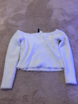 Vit fluffig långärmad tröja - Mysig vit fluffig tröja med lång ärm och croppad passform. Tröjan har en bred halsringning och är gjord i ett supermjukt material som känns skönt mot huden. Perfekt för dig som gillar en enkel men trendig look.