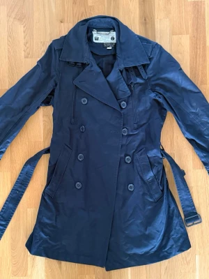 Mörkblå trenchcoat från Diesel - Stilren mörkblå trenchcoat från Diesel med dubbelknäppning, klassisk krage och avtagbart midjebälte. Jackan har två snedställda fickor framtill och justerbara spännen vid ärmsluten. Perfekt för en trendig och tidlös look.