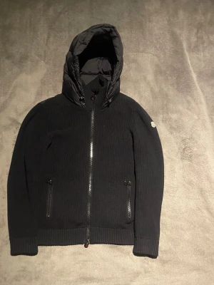 Moncler cardigan  - Snygg svart dunjacka från Moncler med stickade ärmar och quiltad framsida och rygg. Jackan har två dragkedjefickor fram, hög krage och en stor vadderad huva. Moncler-logga på vänster ärm. Perfekt för kalla vinterdagar.