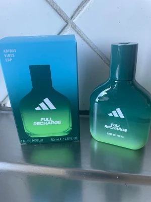 Adidas Full Recharge Eau de Parfum - Adidas Full Recharge Eau de Parfum, 50 ml, med en snygg grön-blå glasflaska och matchande förpackning. Doften är fräsch och energigivande, perfekt för dig som gillar sportiga och moderna parfymer. Flaskan har en stilren design med Adidas-logga.