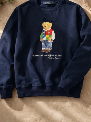 Mörkblå sweatshirt Polo Bear - Cool mörkblå sweatshirt från Ralph Lauren med Polo Bear-motiv på bröstet. Tröjan har rund hals och ribbade muddar. Materialet är mjukt och bekvämt, perfekt för en avslappnad look. Motiv med björn i färgglad outfit och texten 'POLO BEAR by RALPH LAUREN'.