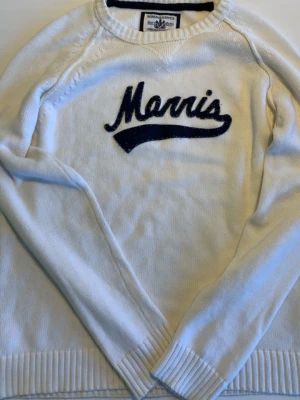 Vit stickad tröja från Morris - Vit stickad tröja från Morris med blå broderad logga på bröstet. Tröjan har rund halsringning, långa ärmar och ribbade muddar vid ärmslut och nederkant. Perfekt för en avslappnad och stilren look.