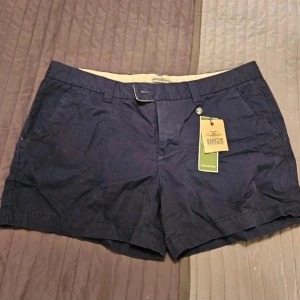 Shorts Hampton Republic 46 - Snygga marinblå shorts från Hampton Republic med klassisk passform, bälteshällor och sidofickor. Perfekta för sommaren och har en stilren look med knapp och dragkedja framtill. Materialet känns mjukt och bekvämt.  Kan även hämtas I Partille  E5