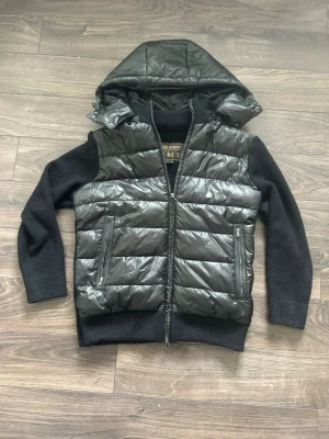 Svart hybridjacka från Leon Marcél - Svart cardiganjacka från Leon Marcél med quiltad kropp i blank polyester och stickade ärmar i 100% bomull. Jackan har borttagbar huva, dragkedja framtill och två fickor med dragkedja. Knappt använd. Nypris hos Leon Marcél: 1400kr. Mitt pris: 899kr