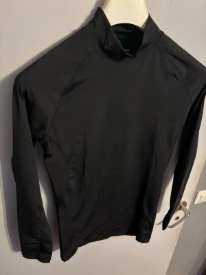 Svart långärmad träningströja Under Armour - Säljer en svart långärmad träningströja från Under Armour med hög krage och logotyp på bröstet. Tröjan är tillverkad i ett stretchigt och fukttransporterande material som passar perfekt för sport och träning.