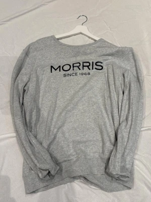 Grå sweatshirt från Morris - Säljer en stilren grå sweatshirt från Morris med svart logotyp och texten 'SINCE 1968' på bröstet. Tröjan har rund halsringning och långa ärmar. Perfekt för en avslappnad och trendig look.