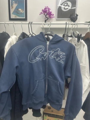 Blå Croxx zip hoodie med tryck - Mörkblå hoodie med dragkedja från Cortiez. Stor vit logga Klassisk huva och ribbade muddar vid ärmar och midja. Perfekt för en chill och street-inspirerad look. NY PRIS 1500kr