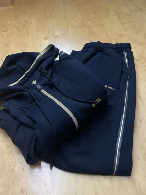 Mörkblå BOSS tracksuit med gulddetaljer - Snygg mörkblå tracksuit från BOSS med guldfärgade detaljer och dragkedja på hoodien. Setet består av en zip-hoodie med ribbade muddar och matchande byxor med guldrand längs sidorna. Perfekt för chill dagar eller när du vill vara bekväm men ändå se fräsch ut.