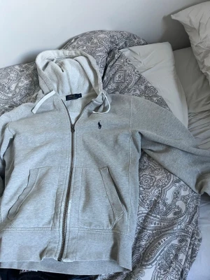 Grå zip hoodie från Polo Ralph Lauren - Snygg grå hoodie med dragkedja från Polo Ralph Lauren. Klassisk design med huva och vita dragsnören, samt två stora fickor framtill. Ikoniska mörkblå loggan broderad på bröstet. Perfekt för en chill och stilren look.