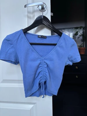 Blå croppad topp från Zara - Blå croppad topp från Zara med ribbad struktur och korta ärmar. Toppen har en djup V-ringning och snygg rynkdetalj med knyt framtill. 
