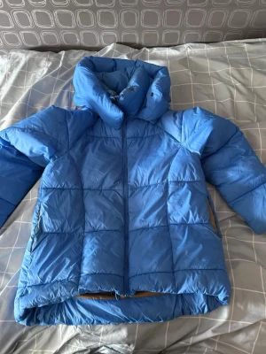 Blå pufferjacka med hög krage - Snygg blå pufferjacka med hög krage och dragkedja framtill. Jackan har quiltad design och två sidofickor med dragkedja. Perfekt för kalla dagar när du vill hålla dig varm och se cool ut.