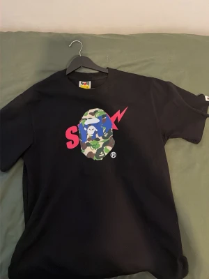 Svart BAPE t-shirt med camo-tryck - Säljer en svart t-shirt från BAPE med ikoniskt camo-tryck och färgglad grafik på bröstet. Klassisk passform och rund hals. Perfekt för dig som gillar streetwear och vill sticka ut med en unik design.