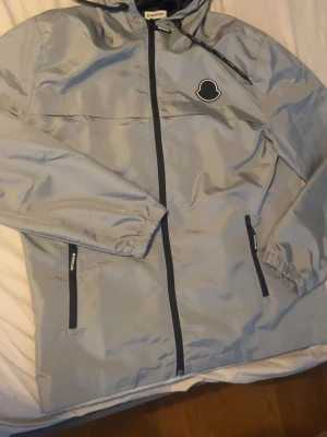 Ljusgrå vindjacka från Moncler - Snygg ljusgrå vindjacka från Moncler med svart dragkedja och detaljer. Jackan har huva, två fickor med dragkedja och ett stilrent Moncler-märke på bröstet. Tillverkad i lätt och glansigt material som passar perfekt till streetwear.
