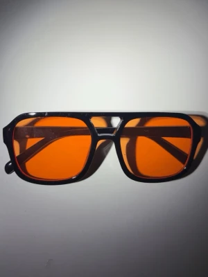 Svarta solglasögon med orange glas - Snygga solglasögon med svart, kantig båge och rektangulära orangefärgade glas. Glasögonen har en retroinspirerad design och passar perfekt för dig som vill sticka ut med en cool accessoar. Tillverkade i syntetmaterial.