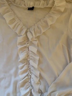 Vit blus från Gina Tricot - Söt vit blus från Gina Tricot med volangdetaljer längs knäppningen och runt halsringningen. Blusen har v-ringning och knappar framtill. 