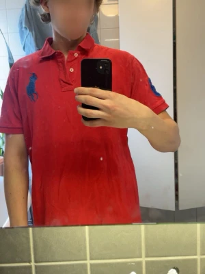 Röd pikétröja från Ralph Lauren - Klassisk röd pikétröja från Ralph Lauren med blå logga på bröstet och siffran 3 på ärmen. Tröjan har krage, knappar vid halsen och korta ärmar. Perfekt för en avslappnad men ändå stilren look.