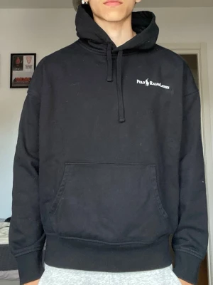 Svart hoodie från Polo Ralph Lauren - Svart hoodie från Polo Ralph Lauren med vit logga på bröstet. Klassisk känguruficka framtill, dragsko i huvan och relaxed fit. Perfekt för en chill och stilren look. Tillverkad i mjukt bomullsmaterial som är skönt mot huden.