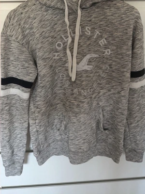 Gråmelerad hoodie från Hollister - Snygg gråmelerad hoodie från Hollister med vit logga och text på bröstet. Har huva med vita snören och känguruficka framtill. Svarta och vita ränder på ärmarna ger en sportig vibe. Perfekt för chill dagar eller när du vill ha en avslappnad stil.
