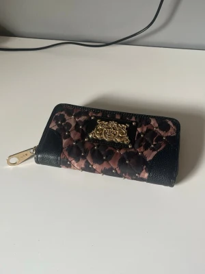 Juicy Couture leopardmönstrad plånbok - Säljer en snygg plånbok från Juicy Couture med leopardmönster i rosa och svart, guldfärgade detaljer och quiltad design. Plånboken har dragkedja runtom, ett guldfärgat emblem framtill och extra fack med dragkedja på baksidan. Perfekt accessoar för dig som gillar statement-plånböcker.