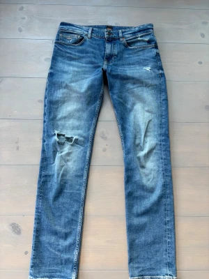 HUGO BOSS DELAWARE JEANS - Snygga blå slim fit jeans från Delaware med slitningar och hål på vänster knä. Klassisk femficksmodell med orangea sömmar och dragkedjegylf. Perfekta för en avslappnad och trendig look.