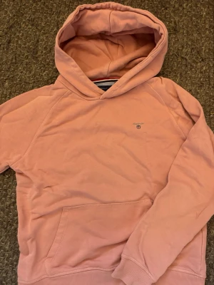 Rosa hoodie från GANT - Säljer en snygg rosa hoodie från GANT med broderad logga på bröstet. Hoodien har en klassisk känguruficka framtill, ribbade muddar och en mjuk insida. Perfekt för dig som gillar stilrena och bekväma plagg.