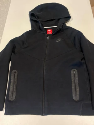 Svart hoodie med dragkedja från Nike - Snygg svart hoodie från Nike med hel dragkedja framtill och två fickor med dragkedja. Tröjan har huva och diskret Nike-logga på bröstet. Perfekt för en sportig och avslappnad stil.