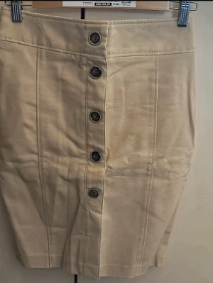 Beige pennkjol med knappar från Zara - Stilren beige pennkjol med knappar framtill och dekorativa fickor bak. Kjolen har en smal passform och är tillverkad i ett mjukt bomullsmaterial. Perfekt för dig som gillar klassisk och enkel stil.
