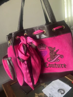 Juicy Couture rosa handväska - Juicy Couture handväska i starkt rosa mocka med bruna detaljer i konstläder. Väskan har broderade loggor, guldfärgade metalldetaljer och en matchande rosa scarf knuten på handtaget. Praktisk storlek med innerfack och dekorativt hjärta med 'J' på baksidan.