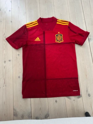 Spansk landslagströja Adidas röd - Säljer en röd spansk landslagströja från Adidas med gula detaljer och tre ränder på axlarna. Tröjan har broderad landslagssköld och Adidas-logga på bröstet. Perfekt för fotbollsfans som vill stötta Spanien på match eller träning.