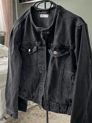 Svart jeansjacka från Ginatricot - Stilren svart jeansjacka från Ginatricot med raka linjer. Jackan har två dekorfickor med lock och silverfärgade knappar framtill. Tillverkad i robust jeansmaterial och har en kraglös halsringning. Inte använd men utan prislapp.
