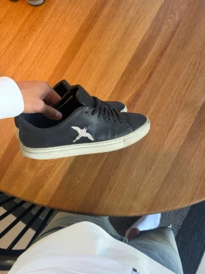 Svarta Axel Arigato sneakers - Säljer ett par svarta sneakers från Axel Arigato med vit fågelbrodyr på sidan och vit sula. Skorna har klassisk låg profil, svarta skosnören och är tillverkade i slätt läder. Perfekta för dig som gillar stilrena och trendiga sneakers.