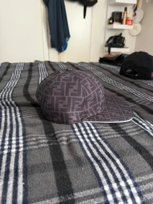 Mörkgrå Fendi keps med FF-mönster - Säljer en snygg mörkgrå keps från Fendi med det klassiska FF-logomönstret över hela kepsen. Kepsen har böjd skärm och justerbar rem bak. Materialet känns slitstarkt och kepsen har en modern streetwear-vibe. Perfekt för dig som gillar exklusiva accessoarer.