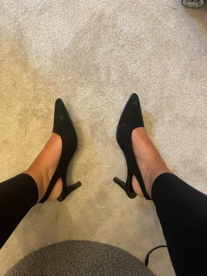 Svarta spetsiga pumps med smal klack - Säljer ett par klassiska svarta pumps med spetsig tå och smal klack. Skorna har en elegant siluett och är tillverkade i ett material som ser ut som mocka. Perfekta för dig som vill ha en stilren och tidlös look.