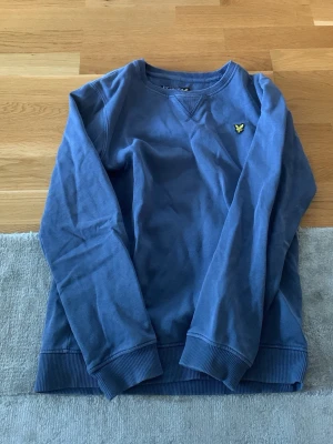 Blå sweatshirt från Lyle & Scott - Blå sweatshirt från Lyle & Scott med rund halsringning och gul broderad logga på bröstet. Tröjan har ribbade muddar vid ärmslut och nederkant samt en relaxed passform. Perfekt för en chill och stilren look. Är en 165cm och passar helt ok och är i mycket bra skick