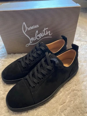 Svarta sneakers från Christian Louboutin - Stilrena svarta sneakers i mocka från Christian Louboutin med klassisk röd sula och svarta skosnören. Låg modell med diskret logga på sulan och insidan. Perfekta för dig som vill ha en lyxig och tidlös look. Otroligt bra skick inte mer använda än enstaka gånger.