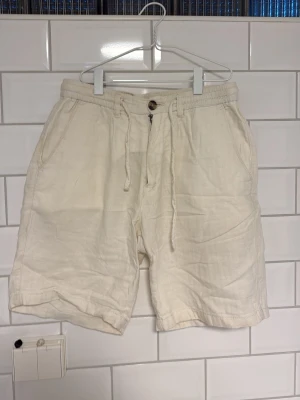 Vita linneshorts med snörning - Säljer ett par vita shorts i linne med snörning i midjan. De har två sidofickor fram och två bakfickor med knapp. Perfekta för varma dagar och har en avslappnad passform.