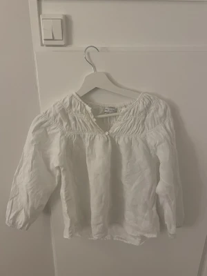 Vit blus med smock från Zara - Söt vit blus från Zara med smockdetaljer vid axlar och halsringning. Blusen har knappar framtill och är tillverkad i ett lätt, luftigt tyg. Perfekt till jeans eller kjol för en fräsch look.