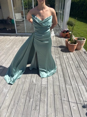 Ljusgrön långklänning med slits - Elegant ljusgrön långklänning i glansigt tyg med draperad omlottringning och hög slits framtill. Klänningen är axelbandslös och har ett långt släp som ger en dramatisk look. Perfekt för bal eller festliga tillfällen. I storlek 36 men går att sy in