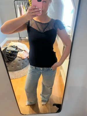 Svart topp med spetsdetaljer - Snygg svart topp med transparent spets upptill och på ärmarna. Toppen har korta ärmar och en rund halsringning med fina spetsmönster som ger en elegant touch. Perfekt att styla med jeans eller kjol.