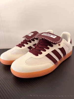 Adidas sneakers med vinröda detaljer - Snygga Adidas sneakers med vinröda läderdetaljer, klassiska tre ränder och vit mocka. Skorna har en unik design med kontrastsömmar på tungan och hälen samt en gummisula i orange. Perfekta för dig som vill sticka ut med stil.