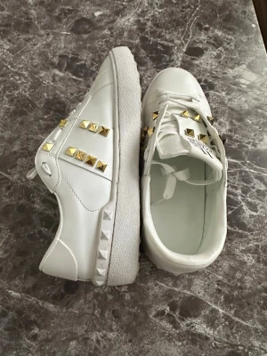 Vita sneakers med guldnitar - Säljer ett par vita sneakers med coola guldiga pyramidnitar på sidorna och över snörningen. Skorna har klassisk låg profil och är tillverkade i slätt syntetmaterial med vit gummisula. Perfekt för dig som vill sticka ut med detaljer.