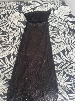 Svart paljettklänning axelbandslös - Säljer en svart, axelbandslös långklänning täckt med glittriga paljetter. Klänningen har en elegant slits på sidan och en figurnära passform som ger en snygg siluett. Perfekt för dig som vill sticka ut på nästa stora event!