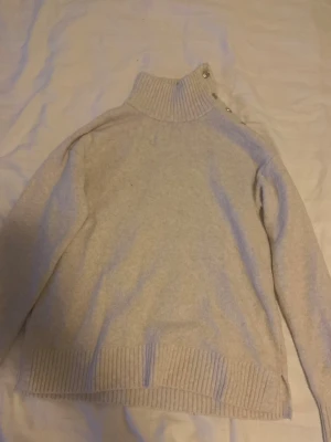 Beige stickad polotröja med knappar - Mysig beige stickad polotröja med ribbad hög krage och dekorativa knappar på axeln. Tröjan har långa ärmar och bred ribbstickning vid ärmslut och nederkant. Perfekt för kalla dagar och enkel att matcha med jeans eller kjol.
