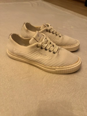 VitBeige sneakers från Gant - Snygga vitbeige sneakers från Gant med ribbad textilovandel och vita gummisulor. Klassisk låg modell med snörning och diskret logga på plösen. Perfekta för en clean och stilren look. Skorna har metallöljetter och en bekväm passform. Storlek 41, men passar 42