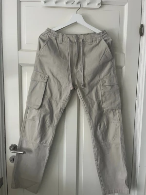 Beige cargobyxor  - Snygga beige cargobyxor från New Garments med stora fickor på sidorna och dragsko i midjan. Byxorna har raka ben och är tillverkade i ett slitstarkt bomullsmaterial. Perfekta för en avslappnad och trendig look.