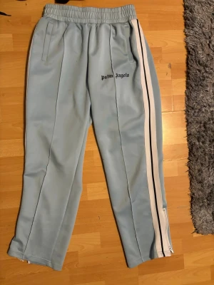 Palm Angels ljusblå mjukisbyxor - Palm Angels ljusblå mjukisbyxor med vita och svarta sidoränder. Byxorna har resår i midjan, raka ben och fickor på sidorna. Märkets logga är broderad i svart på vänster lår. Perfekta för en chill och trendig look.